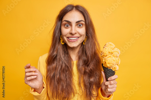 Εκτύπωση καμβά Studio shot of beautiful surprised brunette young woman holds delicious ice cream smiles broadly enjoys eating delicious gelato poses indoor against yellow background