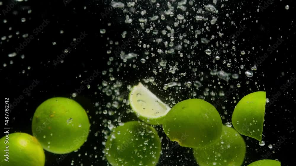 Close-up of falling ripe limes into the sparkling water on black background, making a cocktail of citrus fruits, drinking cold lemonade, shooting of carbonated water with floating sliced fruits. 