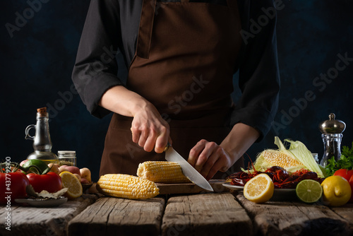 In the photo, the chef is cutting corn into pieces on a cutting board. There are also many ingredients on the wooden table. The cook is wearing a brown apron. Dark background bright paint ingredients photo
