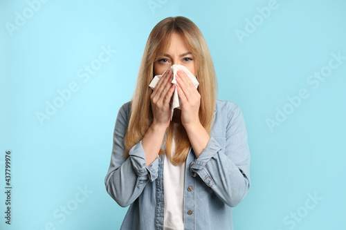 Young woman with tissue suffering from runny nose on light blue background