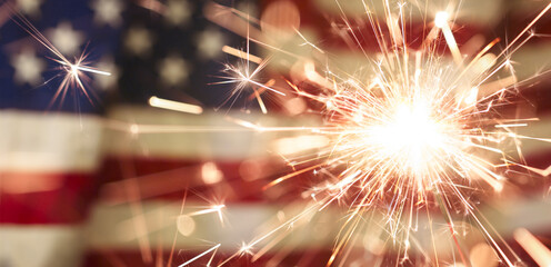 Sparks flying off a burning sparkler in front of the US American flag for patriotic 4th of July celebration.