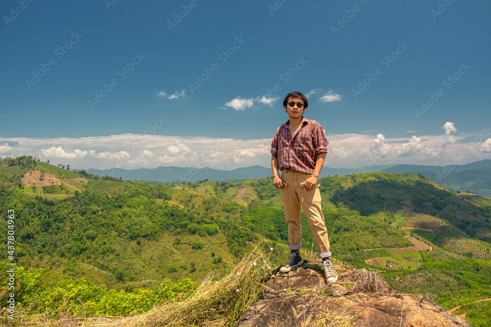Asian man standing on a rock against a backdrop of mountains and sky. A 35-year-old man wearing a striped shirt and brown trousers with sunglasses stands on a rock. Asian man standing on a rock against a backdrop of mountains and sky. A 35-year-old man wearing a striped shirt and brown trousers with sunglasses stands on a rock.