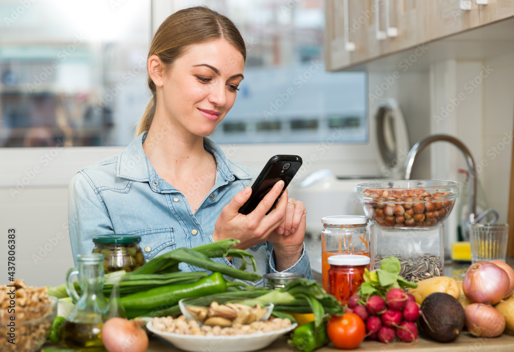 Portrait of glad woman looking at mobile phone while making dish on kitchen at home. High quality photo