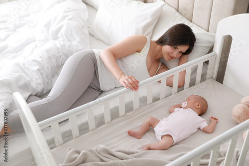 Happy young mother near crib with sleeping newborn baby in bedroom