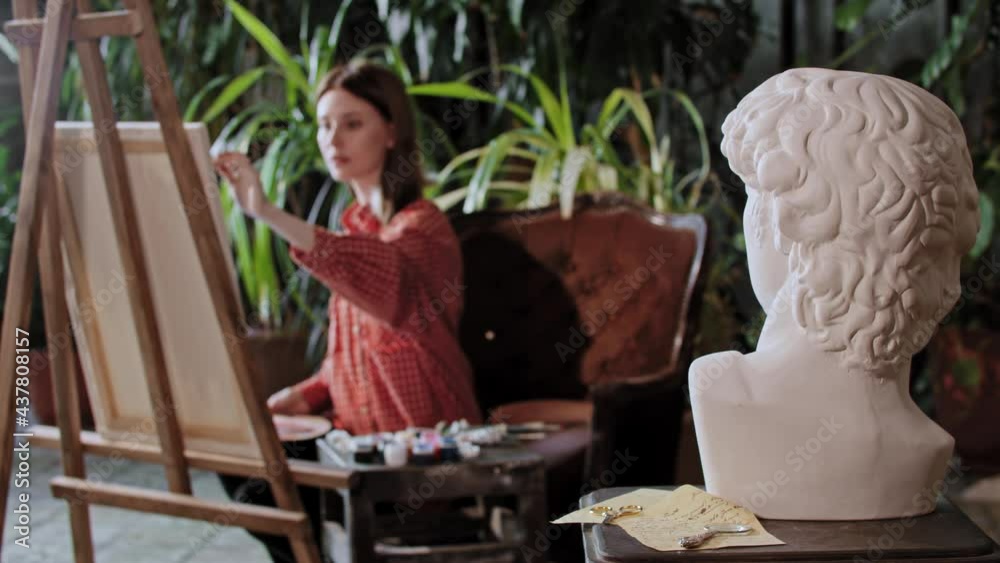 Artist in an art studio with plants - young woman drawing a greek head sculpture on a foreground Artist in an art studio with plants - young woman drawing a greek head sculpture on a foreground