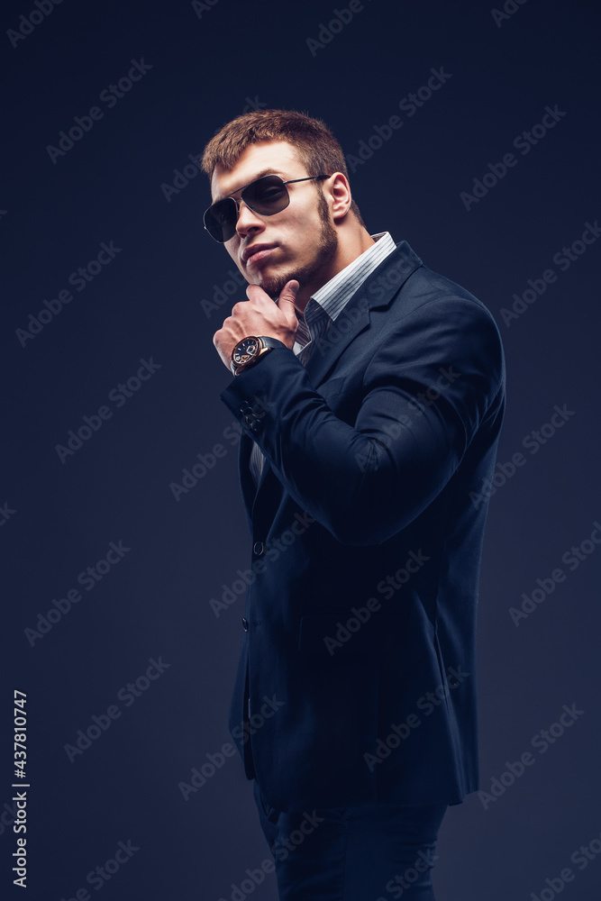 Side view of a Fashion bearded young serious man in sunglasses, luxury suit with one hand in pocket, other is on the chin looking at camera Side view of a Fashion bearded young serious man in sunglasses, luxury suit with one hand in pocket, other is on the chin looking at camera