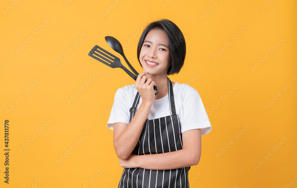 Happy young asian woman wear on an apron and hands showing spatula with pot on light yellow background. Happy young asian woman wear on an apron and hands showing spatula with pot on light yellow background.