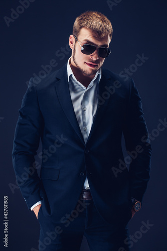 Fashion bearded young serious man in sunglasses, luxury suit looking at camera, hands in pockets 
