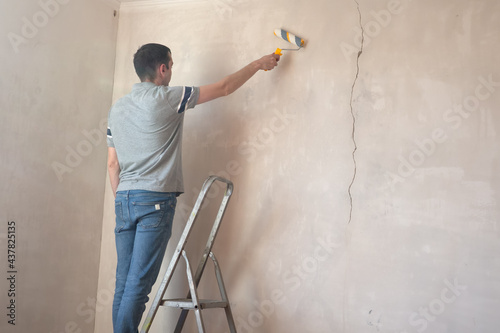 Man priming the wall with a roller. Repair of the interior. Young male decorator painting a wall in the empty room. Concept renovation do it yourself.