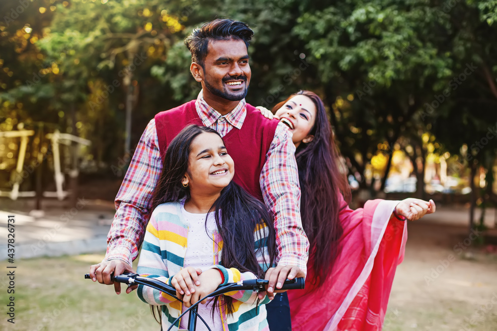 Fototapeta premium Beautiful Indian family - mother, father, daughter - riding a bicycle all together in the park