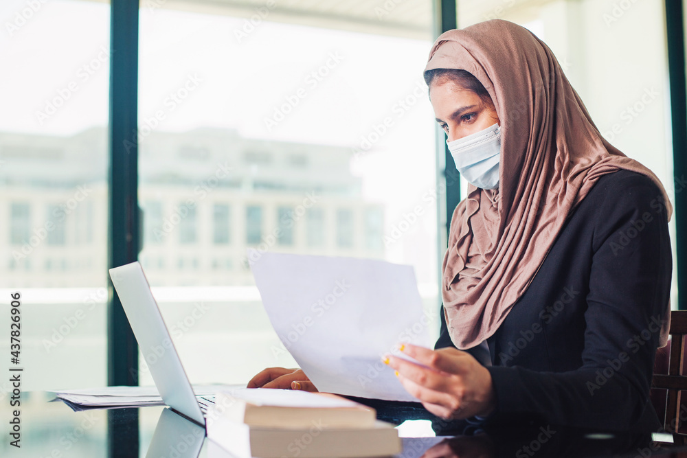 Arabic Muslim woman in hijab and a mask working on her laptop, holding documents