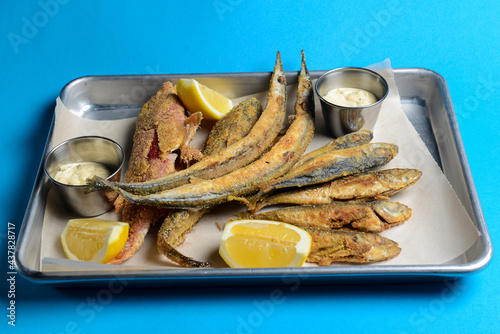 Fried fish with fresh herbs and lemon served on a metal tray over blue background. photo