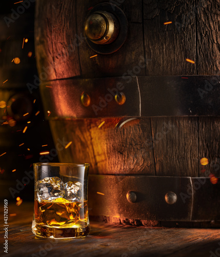 Glass of whisky with ice cubes and old wooden cask at the background.
