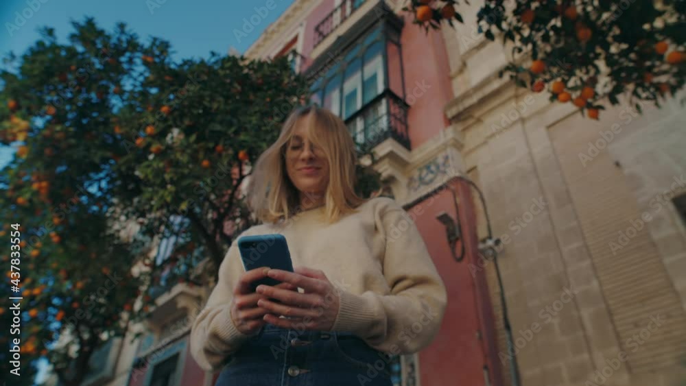 Low angle shot of beautiful young woman text with friends on smartphone. Cinematic shot of attractive female on city street. Millennial use phone, to check social media and do online shopping