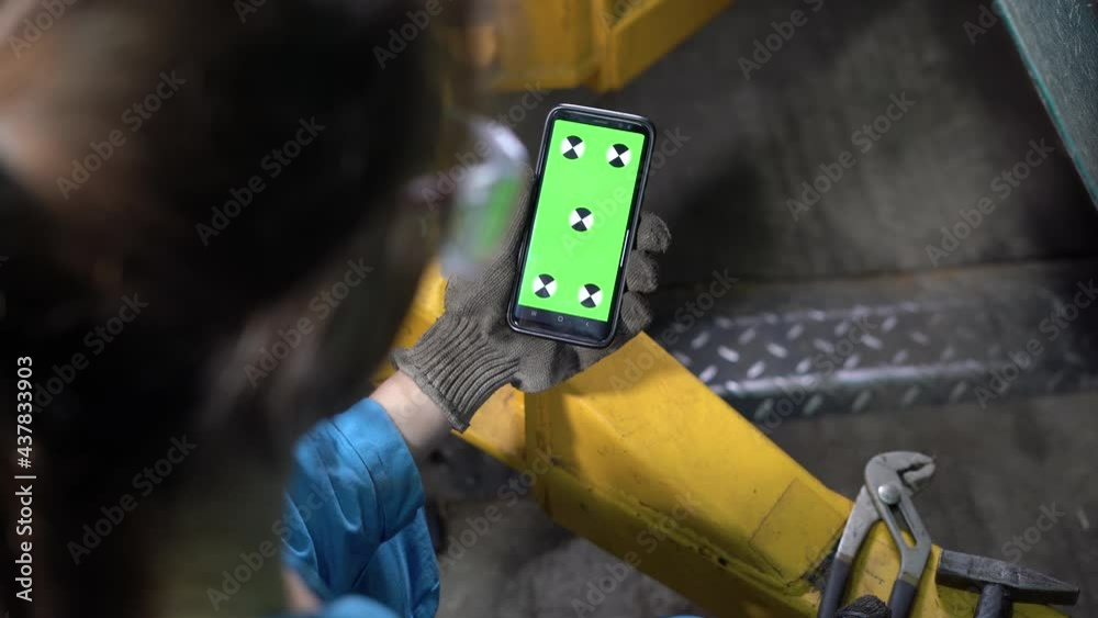 Shoulder view of a woman car mechanic in glasses and overalls holding a smartphone in her hand and looking at a green screen with a chroma key. Video material. Mobile phone.