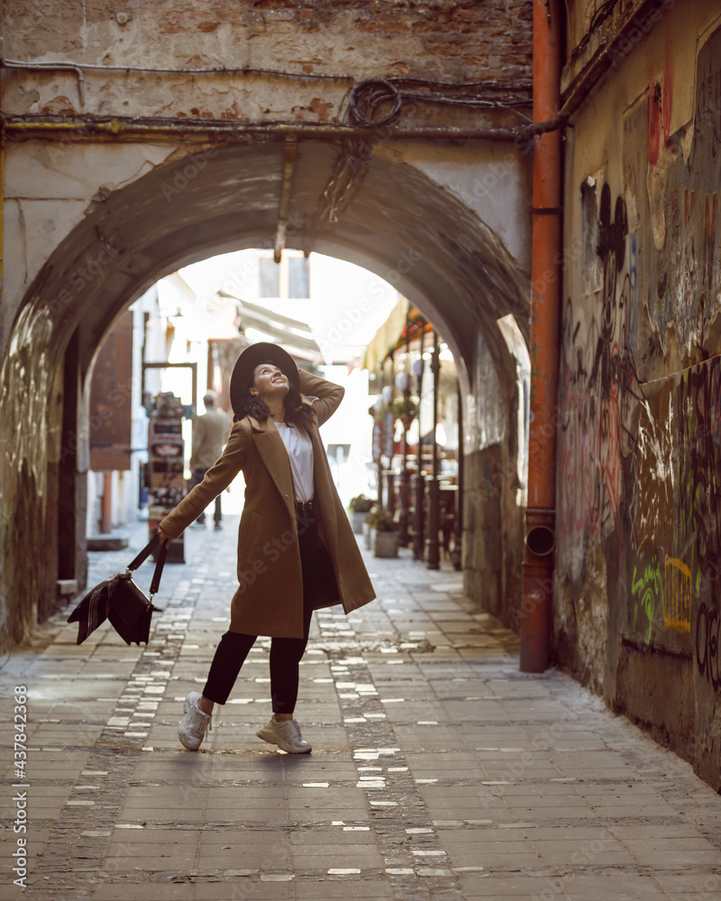 woman in stylish coat and hat walking by small european street