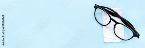 Black rimmed glasses and cleaning cloth on blue background, banner with copy space. White soft tissue for rubbing and polish optics from dirt and dust. Vision correction, ophthalmology, optometry
