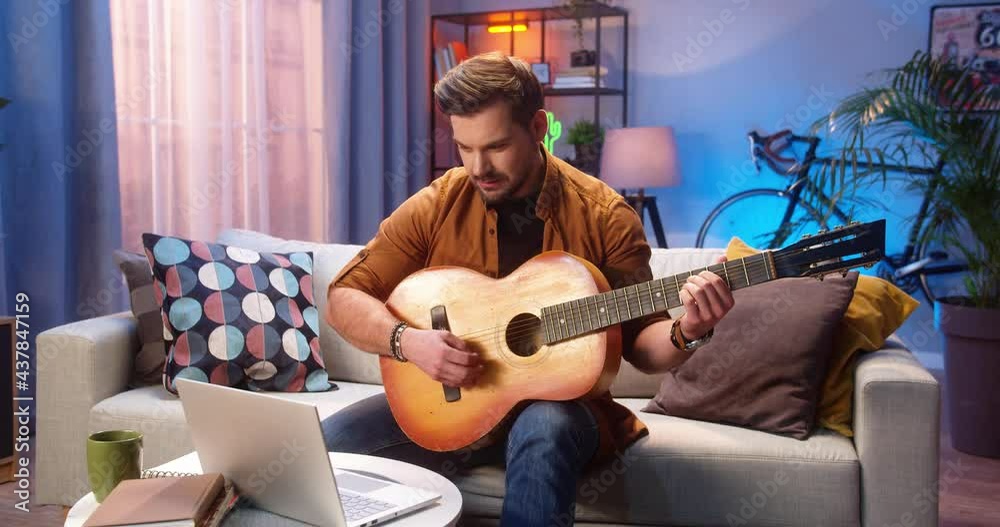 Joyful young Caucasian bearded man having online video lesson on laptop computer learning how to play the guitar while sitting on couch in living room, musician playing the instrument at home