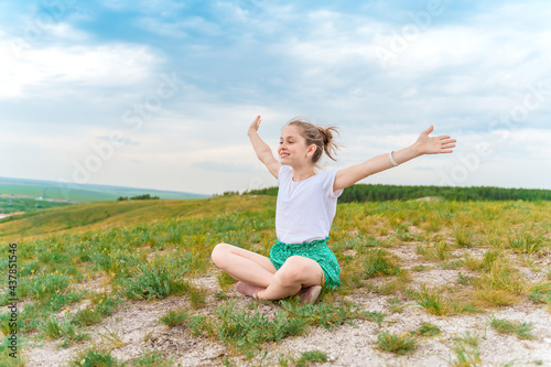 A charming little girl sits on the grass and cheerfully raises her hands against the background of a green meadow and hills