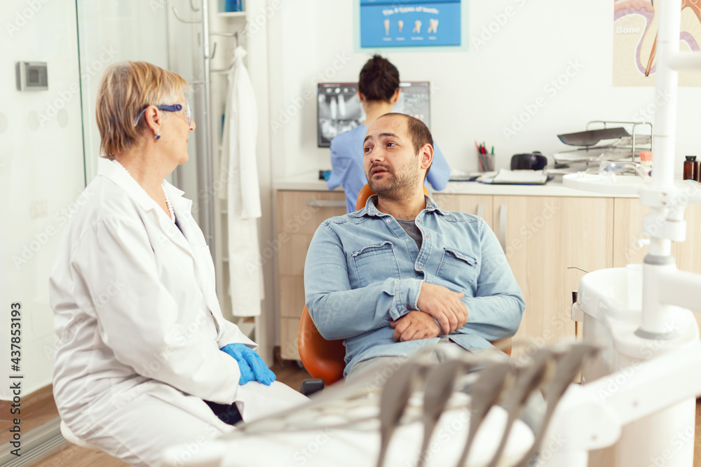 Specialist senior dentist discussing with man patient sitting on dental chair preparing for tooth surgery. In background medical assistant looking into monitor examining stomatological radiography