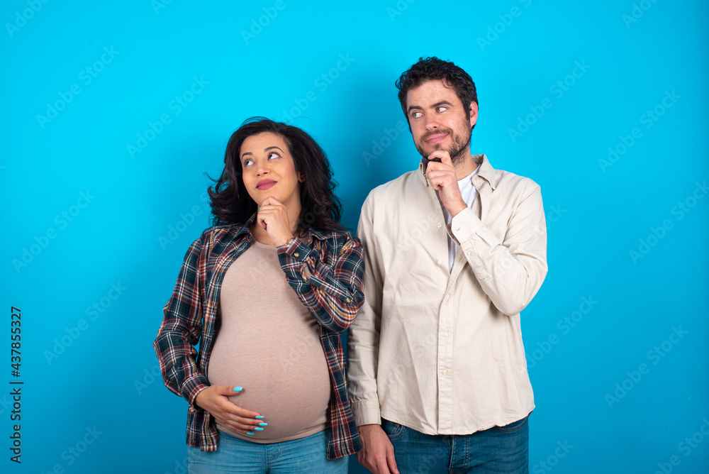 Naklejka premium Thoughtful young couple expecting a baby standing against blue background holds chin and looks away pensively makes up great plan