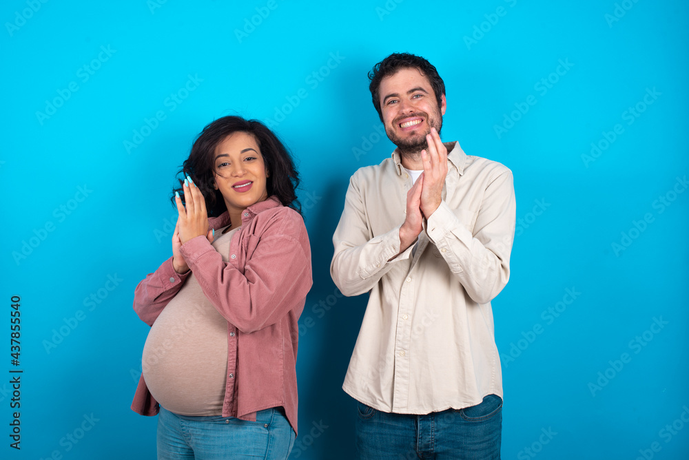 young couple expecting a baby standing against blue background clapping and applauding happy and joyful, smiling proud hands together. young couple expecting a baby standing against blue background clapping and applauding happy and joyful, smiling proud hands together.