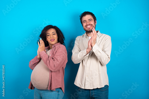 young couple expecting a baby standing against blue background clapping and applauding happy and joyful, smiling proud hands together.