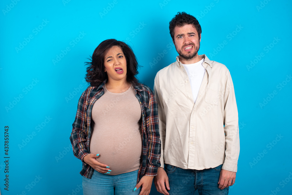 Portrait of dissatisfied young couple expecting a baby standing against blue background smirks face, purses lips and looks with annoyance at camera, discontent hearing something unpleasant Portrait of dissatisfied young couple expecting a baby standing against blue background smirks face, purses lips and looks with annoyance at camera, discontent hearing something unpleasant