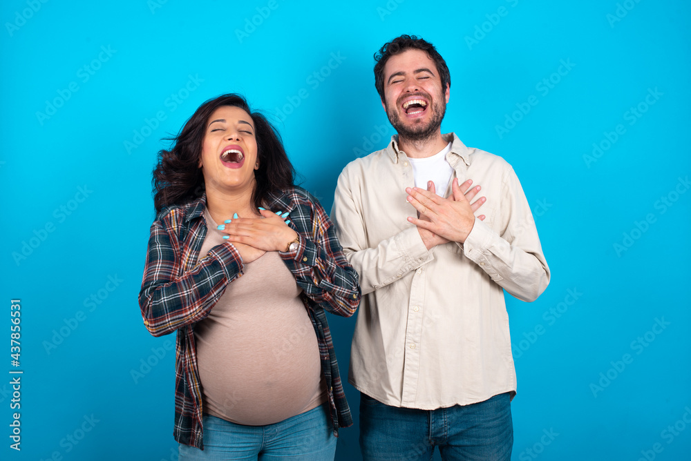 Fototapeta premium young couple expecting a baby standing against blue background expresses happines, laughs pleasantly, keeps hands on heart