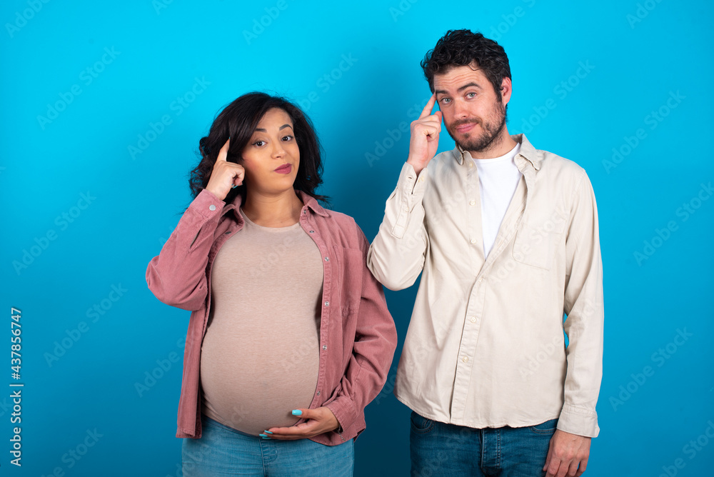 Naklejka premium young couple expecting a baby standing against blue background tries to memorize something, keeps fore finger on temple, reminds information for exam,