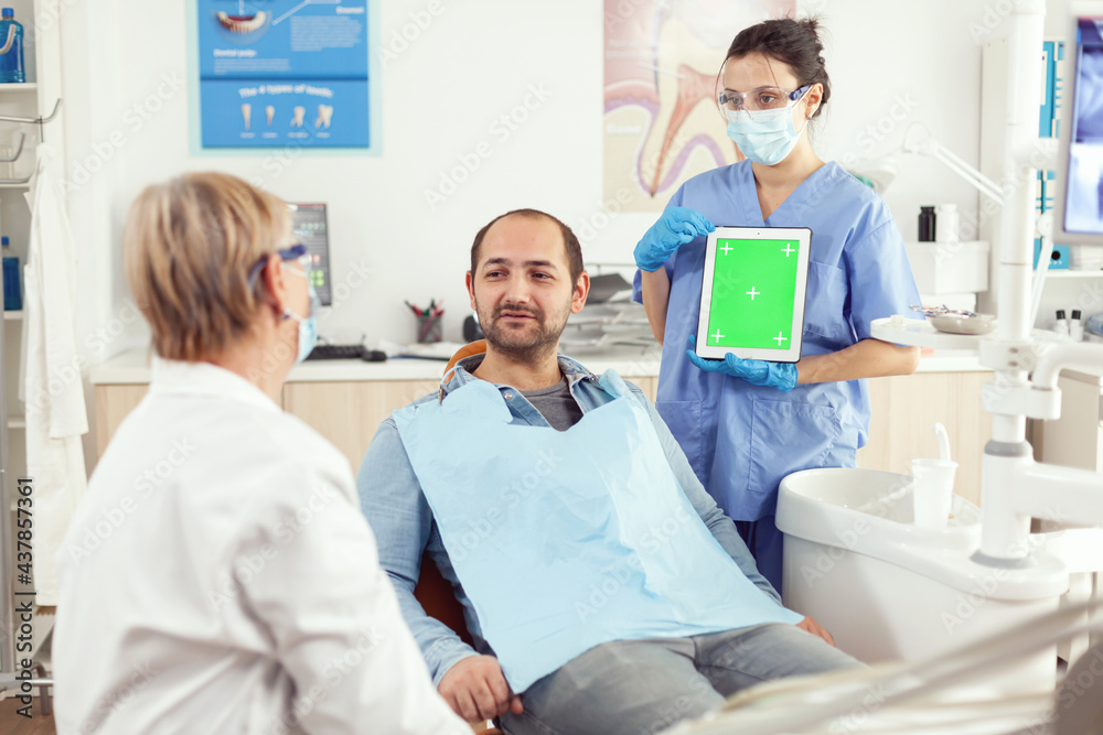 Obraz premium Nurse dentist showing green screen display to stomatology senior doctor while examining tooth pain to man patient sittinh on dental chair. Woman examinating using mock up chroma key tablet