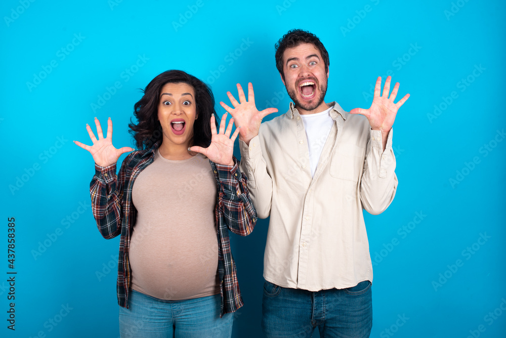 Delighted positive young couple expecting a baby standing against blue background opens mouth and arms palms up after having great result Delighted positive young couple expecting a baby standing against blue background opens mouth and arms palms up after having great result