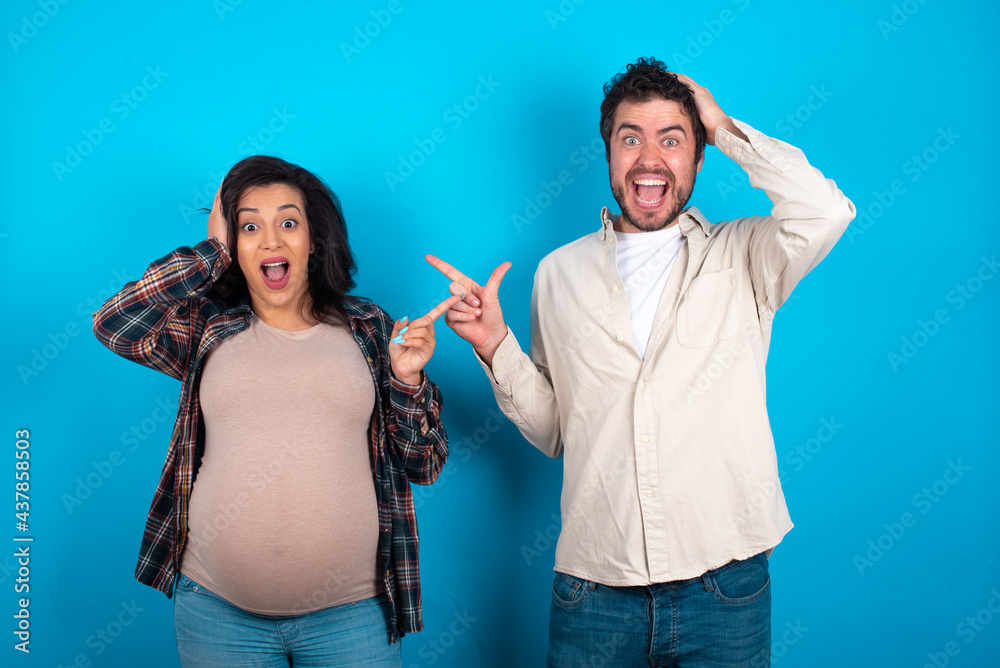 Surprised young couple expecting a baby standing against blue background pointing at empty space holding hand on head Surprised young couple expecting a baby standing against blue background pointing at empty space holding hand on head