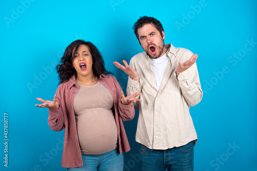 Frustrated young couple expecting a baby standing against blue background feels puzzled and hesitant, shrugs shoulders in bewilderment, keeps mouth widely opened, doesn't know what to do.
