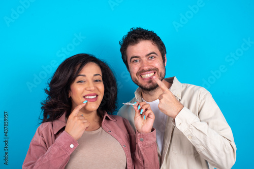 Quadro su tela young couple expecting a baby standing against blue background holding an invisible aligner and pointing to her perfect straight teeth