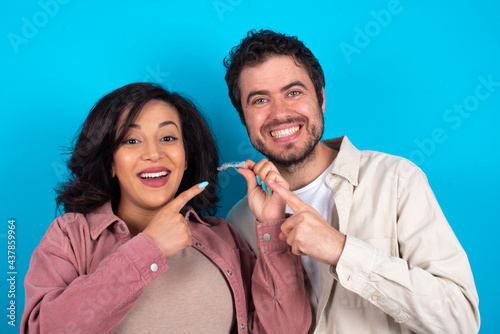 Foto young couple expecting a baby standing against blue background holding an invisible aligner and pointing at it