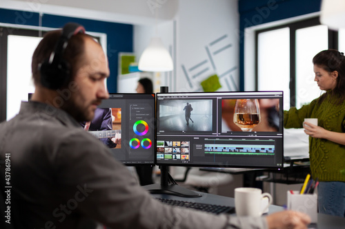 Videographer with headset working with footage and sound on pc with two displays, taking notes while editing customer video. Man processing film montage sitting in creative startup agency office.