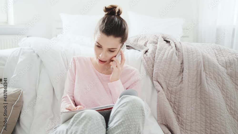 A concentrated girl talking on the phone and writing something while sitting on the floor in a sunny room
