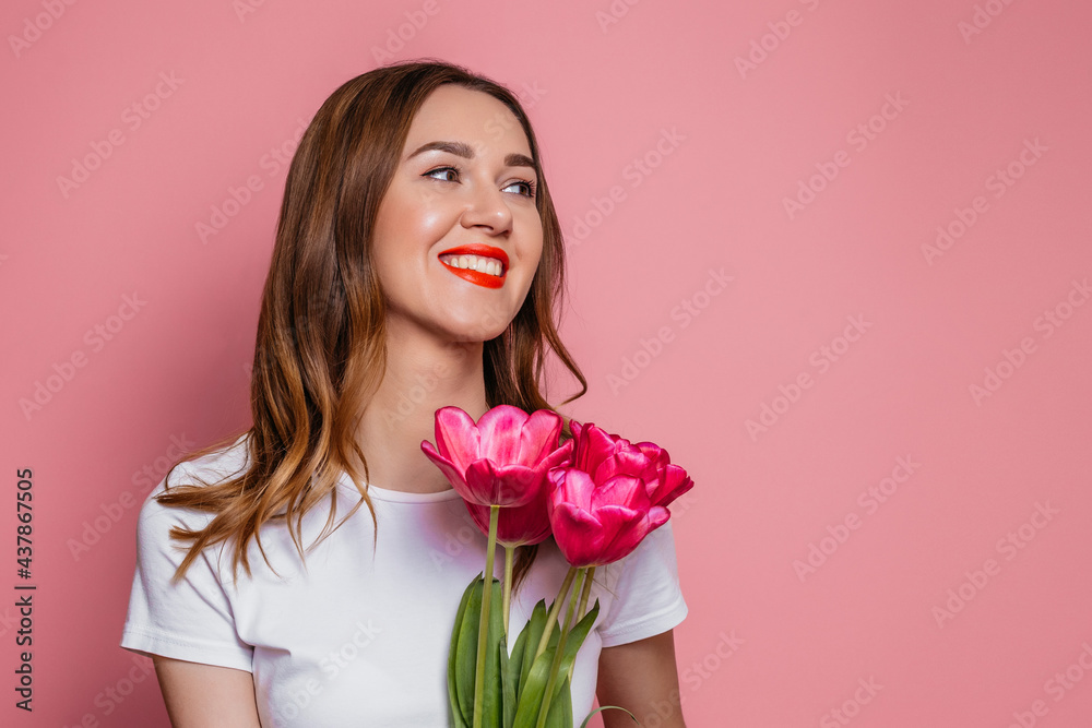 Portrait of a beautiful young woman in a white t-shirt holding big bouquet of tulips isolated over pink background