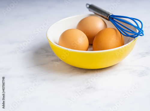 Three brown eggs in a yellow bowl with blue whisk.