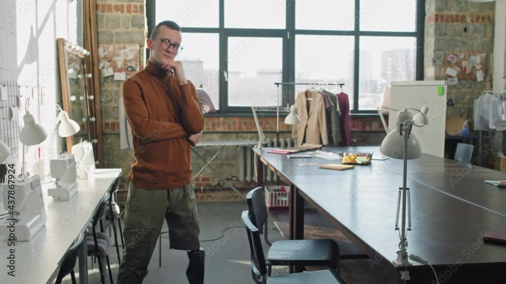 Portrait shot with PAN of young male fashion designer with prosthetic leg standing in airy studio and looking at camera Portrait shot with PAN of young male fashion designer with prosthetic leg standing in airy studio and looking at camera