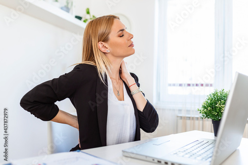 Portrait of young stressed woman sitting at home office desk in front of laptop, touching aching neck with pained expression, suffering from neck pain after working on pc © Graphicroyalty
