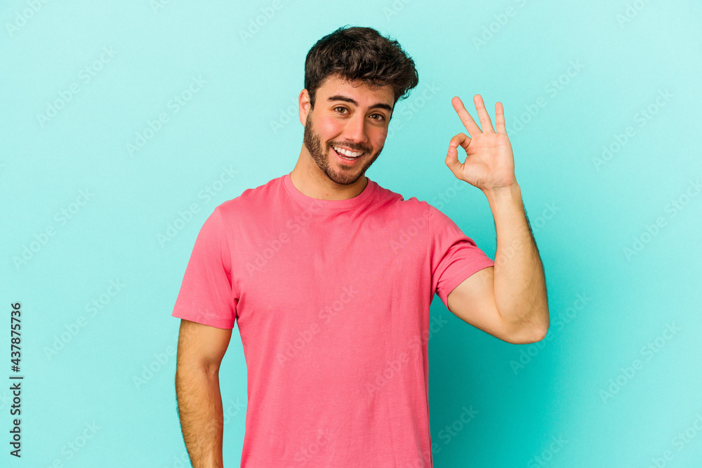 Young caucasian man isolated on blue background winks an eye and holds an okay gesture with hand. Young caucasian man isolated on blue background winks an eye and holds an okay gesture with hand.