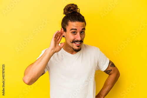Young caucasian man with long hair isolated on yellow background trying to listening a gossip.