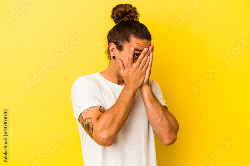 Young caucasian man with long hair isolated on yellow background blink through fingers frightened and nervous.