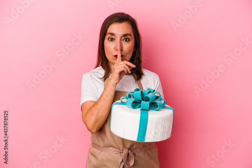 Middle aged pastry chef woman holding a cake isolated on blue background keeping a secret or asking for silence.