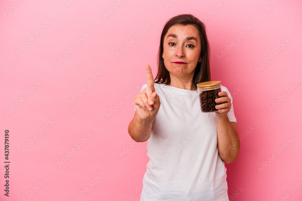 Middle age caucasian woman holding a coffee jar isolated on pink background showing number one with finger. Middle age caucasian woman holding a coffee jar isolated on pink background showing number one with finger.