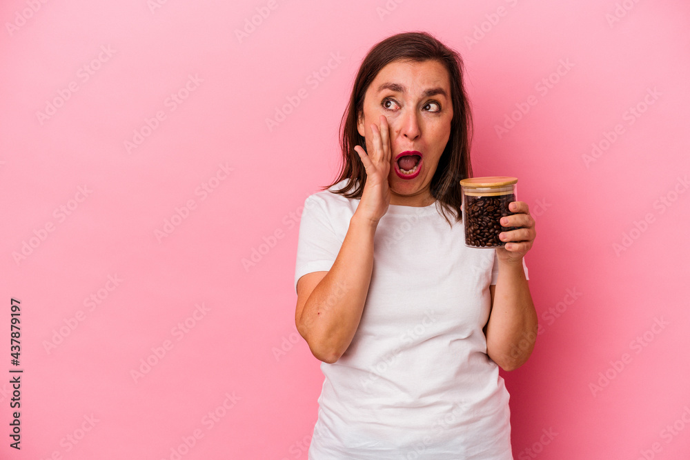 Middle age caucasian woman holding a coffee jar isolated on pink background is saying a secret hot braking news and looking aside