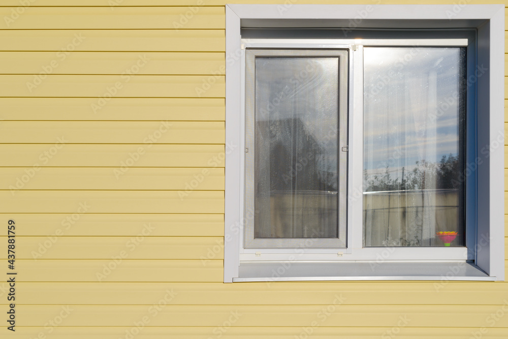 Fototapeta premium The wall of the house is covered with yellow vinyl siding and a plastic white window