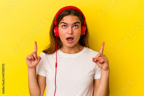 Young caucasian woman listening to music isolated on yellow background pointing upside with opened mouth.